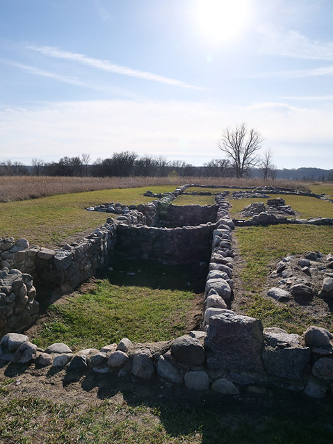 Stone foundation ruins in a grassy field, with trees and sky in the background.
